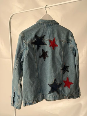 Jeansjacka med stjärnpatches - Jeansjacka med stora dekorativa stjärnpatches i rött och mörkblått på ryggen. Perfekt för dig som vill sticka ut med en unik och trendig look.🩵