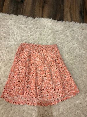 Blommig kortkjol från XSIS - Söt kortkjol från XSIS i en ljus korallrosa färg med små vita och svarta blommor. Kjolen har resår i midjan och är gjord i ett lätt och mjukt material, perfekt för varmare dagar. Passar dig som gillar en avslappnad och feminin stil.