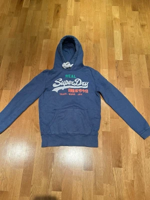 Blå hoodie från Superdry med tryck - Säljer en riktigt schysst Superdry vintage hoodie.                Perfekt för dig med mer avslappnad och snygg stil. Hoodien är i bra skick och har inga märkbara defekter. Har du frågor är det bara att höra av sig!!