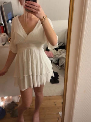 Vit omlottklänning med volang - Söt vit kortklänning med omlott och v-ringning. Klänningen har volangdetaljer och trekvartsärmar, perfekt för en fräsch och somrig look. Tillverkad i ett lätt och luftigt material som känns skönt mot huden.