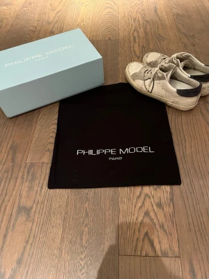  Philippe Model sneakers - Snygga sneakers från Philippe Model i vitt skinn med mörkblå mocka detaljer vid hälen och på sidan. Klassisk låg modell med vita sulor och matchande vita skosnören. Perfekta för en stilren och avslappnad look.