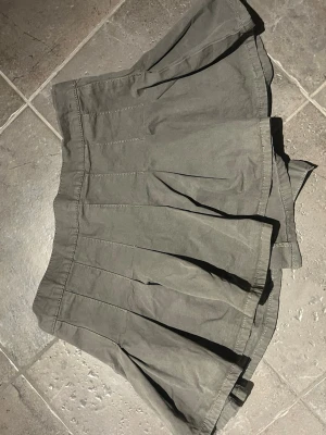 miniskirt med ruffles  - Säljer en grå kortkjol med plisserad design och knappdetalj i midjan. Kjolen har en avslappnad passform och är tillverkad i ett mjukt bomullsliknande material. Perfekt för dig som gillar en enkel men snygg stil.