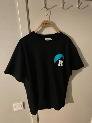 Svart Rhude t-shirt med tryck - Svart t-shirt från Rhude med stort grafiskt tryck i blått och vitt på ryggen och mindre logga på bröstet. Oversized passform och klassisk rund hals. Perfekt för dig som gillar streetwear och vill sticka ut med en unik design.