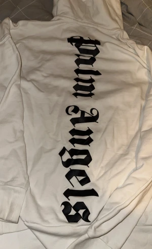 Vit hoodie från Palm Angels - Säljer en vit hoodie från Palm Angels med stor svart logga i gotisk stil på ryggen och mindre text framtill. Hoodien har svart dragsko i huvan och en klassisk magficka. Tillverkad i mjukt bomullsmaterial, perfekt för en avslappnad streetwear-look.