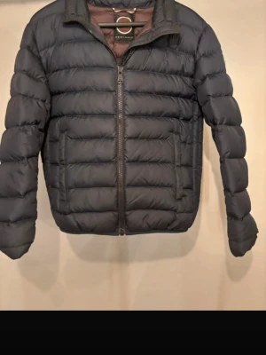 Svart dunjacka från Save The Duck -  Marinblå dunjacka med quiltad design och dragkedja framtill. Jackan har två sidofickor med dragkedja och hög krage. Perfekt för vinterdagar och har en stilren look som passar till det mesta. Storlek 48.