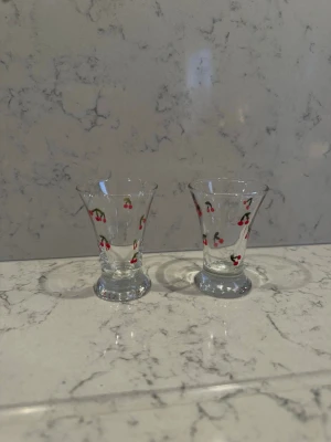 Små glas med körsbärsmotiv - Två små glas i klart glas med sött körsbärsmotiv runt om. Glasen har en lätt utsvängd form och står på en liten fot. Perfekta för shots eller små drinkar och ger en lekfull touch till dukningen.