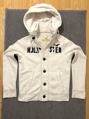 Vintage Hollister Cardigan Med Knappar - Väldigt sällsynt och vintage Hollister hoodie med knappar | Tröjan är i väldigt bra skick utan defekter, handledarna kan uppfattas vara lite större| Längd ≈ 63cm, Bredd armhåla-amrhåla ≈ 53cm | Modell 175cm, 68kg | Hör av vid ytterligare frågor och priset kan diskuteras vid snabbt köp! |