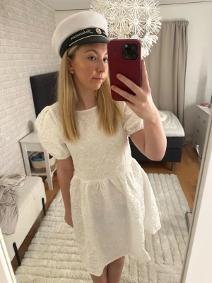 Studentklänning  - Superfin vit kort klänning med puffärmar och strukturmönster. Klänningen har rund halsringning och en lätt utställd kjol. Perfekt för student eller sommarens alla tillfällen.