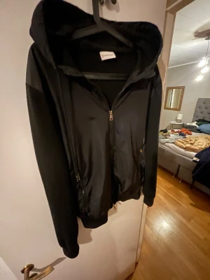 Svart jacka med huva från Moncler - Snygg svart jacka från Moncler med huva och dragkedja framtill. Jackan har stickade ärmar och detaljer samt slät framsida. Märkeslogga på ena ärmen. Perfekt för dig som vill ha en stilren och modern look.