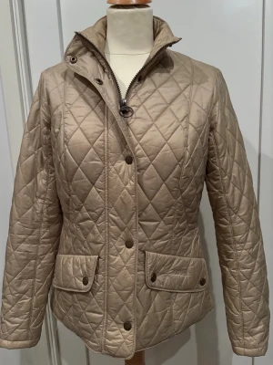 Beige quiltad jacka från Barbour - Stilren beige quiltad jacka från Barbour med tryckknappar och dragkedja framtill. Jackan har två fickor med lock och Barbour-logga, samt hög krage. Perfekt för dig som gillar klassisk och tidlös design.