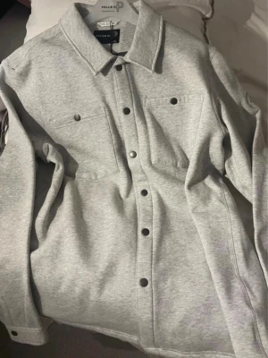 Ljusgrå overshirt från Pelle P - Stilren ljusgrå overshirt från Pelle P med tryckknappar framtill och två bröstfickor. Overshirten har klassisk krage och är tillverkad i ett mjukt sweatshirtliknande material. Perfekt lager på lager-plagg för en avslappnad look.