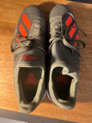 Gråa Adidas fotbollsskor med röda detaljer - Snygga fotbollsskor från Adidas i grått med röda detaljer och klassiska tre ränder på sidan. Skorna har snörning och en ovandel i syntetmaterial som ger bra passform och kontroll på planen. Perfekta för dig som vill sticka ut på fotbollsplanen.