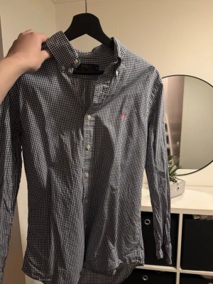 Rutig skjorta från Polo Ralph Lauren - Klassisk rutig skjorta från Polo Ralph Lauren i blått och vitt. Skjortan har lång ärm, knappar framtill och en liten rosa logga broderad på bröstet.