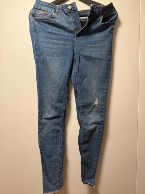 Blå skinny jeans från Pieces - Säljer ett par blå skinny jeans från Pieces med slitning på ena benet och råa kanter vid bensluten. Jeansen har klassisk femficksdesign, hög midja och dragkedjegylf. Perfekta för en avslappnad och trendig look.