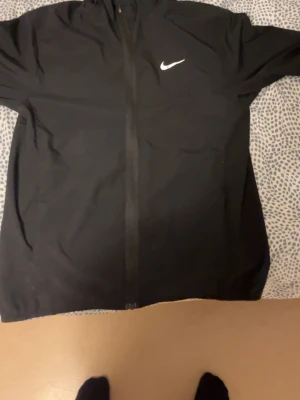 Svart vindjacka från Nike med huva - Säljer en svart vindjacka från Nike med klassisk vit logga på bröstet. Jackan har huva och dragkedja framtill, perfekt för blåsiga dagar. Enkel och stilren design som passar till det mesta.