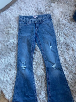 Blå bootcut jeans med slitningar - Säljer ett par blå bootcut jeans med slitningar på benen och snygga broderade detaljer på bakfickorna. Jeansen har låg midja och klassisk femficksmodell. Perfekta för dig som gillar en avslappnad och trendig stil.