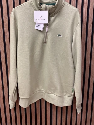 Grön halvzip tröja  - Snygg beige tröja med halv dragkedja Tröjan har hög krage och ribbade muddar vid ärmslut och nederkant. Perfekt för en clean och stilren look.