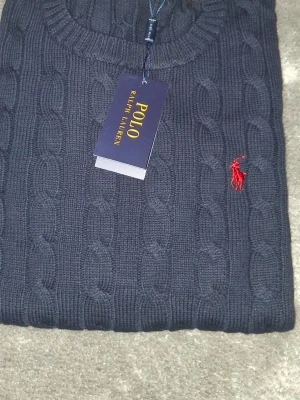 Mörkblå kabelstickad tröja Polo Ralph Lauren - Kabelstickad mörkblå tröja från Polo Ralph Lauren med rund halsringning och klassisk röd broderad logga på bröstet. Tröjan är i mjukt material och har ribbade muddar vid ärmslut och nederkant. Perfekt för en stilren och tidlös look.