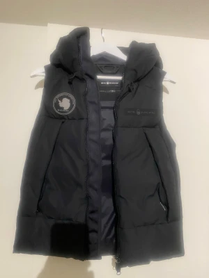 Svart dunväst från Sail Racing - Stilren svart dunväst från Sail Racing med huva och dragkedja framtill. Västen har två fickor med dragkedja och logga på bröstet samt ett märke på vänster sida. Perfekt för lager-på-lager och streetwear.