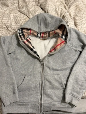 Burberry hoodie  - Snygg grå hoodie med dragkedja från Burberry. Klassisk rutmönstrad insida i huvan och broderad logga på bröstet. Mjuk bomullsblandning, två fickor fram och ribbade muddar. Perfekt för en chill och stilren look.