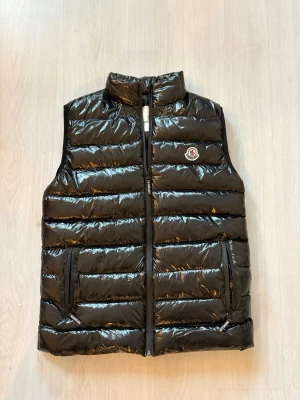 Svart dunväst från Moncler - Helt ny! Dock utan prislapp)Snygg svart dunväst från Moncler med glansig finish och klassisk quiltad design. Västen har hög krage, dragkedja framtill och två fickor med dragkedja. Moncler-logga på bröstet ger en exklusiv touch. Perfekt för lager-på-lager-stil.
