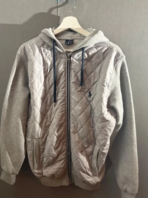 Grå quiltad hoodie från Ralph Lauren - Snygg grå hoodie från Ralph Lauren med quiltad framsida och dragkedja. Har huva med mörka snören och broderad logga på bröstet. Ärmar och baksida i mjuk sweatshirtmaterial, perfekt för en avslappnad men stilren look.