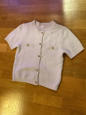 Vit kortärmad kofta med guldknappar - Kortärmad kofta i mjuk, vit stickad kvalitet med ribbade kanter och dekorativa guldknappar framtill samt på två fejkfickor. Klassisk rund halsringning och rak passform. Perfekt för en stilren och trendig look.