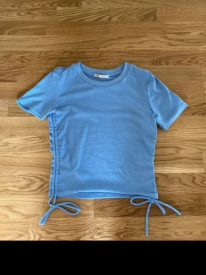 Blå ribbad topp med knyt från Zara - Trendig ljusblå ribbad topp från Zara med korta ärmar och rund halsringning. Snygga knytband i sidan ger en cool detalj och möjlighet att justera passformen. Perfekt till jeans eller kjol för en avslappnad look.