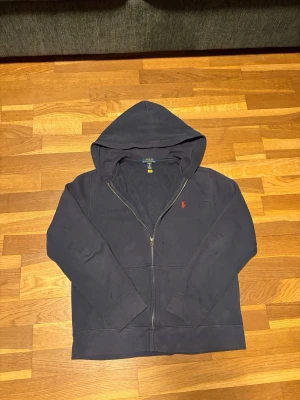 Mörkblå hoodie från Polo Ralph Lauren - Mörkblå hoodie med dragkedja från Polo Ralph Lauren. Klassisk design med huva, två fickor framtill och den ikoniska röda loggan broderad på bröstet. Tillverkad i mjukt bomullsmaterial som är perfekt för en avslappnad stil.