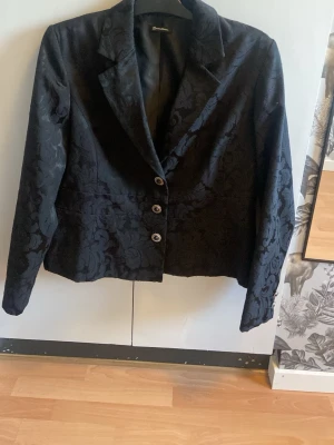 Svart mönstrad kavaj från Bonmarche - Stilren svart kavaj från Bonmarche med diskret blommönster i tyget. Kavajen har tre dekorativa knappar framtill och klassisk krage. Materialet ser ut att vara en blandning med struktur, vilket ger en elegant känsla. Perfekt för dig som vill ha något extra till outfiten.