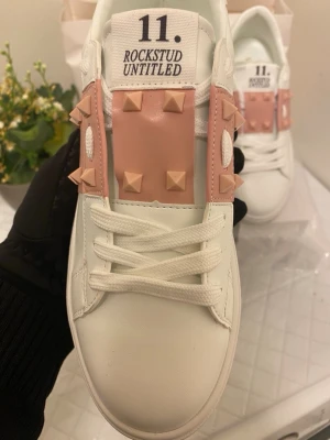 Valentino Rockstud Untitled sneakers - Snygga vita sneakers från Valentino med rosa detaljer och ikoniska nitar på sidorna och över vristen. Skorna har en låg profil, klassiska snörningar och är tillverkade i läder. Perfekta för dig som vill sticka ut med en lyxig och trendig look.