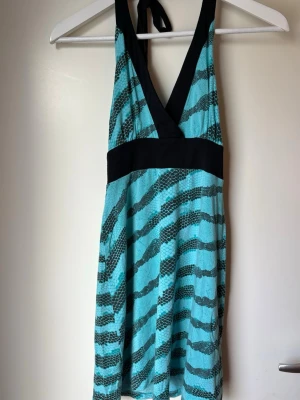 Turkos och svart halterneckklänning - Snygg kortklänning i turkos med svarta zebramönster och bred svart midjedetalj. Klänningen har halterneck med knyt i nacken och v-ringad urringning. Perfekt för dig som vill sticka ut med färg och mönster.
