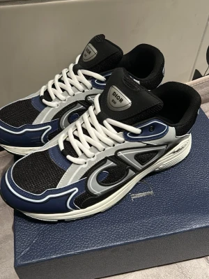Dior B30 sneakers blå och svart - Snygga Dior B30 sneakers i blått, svart och vitt med vita skosnören. Ovandelen är i mesh och läder med stora Dior-loggor på sidan. Tjock sula med svart och vit detaljering. Perfekta för dig som gillar exklusiva och trendiga sneakers.