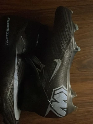 Nike Air Zoom svarta fotbollsskor - Säljer ett par svarta Nike Air Zoom fotbollsskor med silverdetaljer och en snygg logga på hälen. Skorna har dobbar för bra grepp på planen och är tillverkade i syntetmaterial för lätt vikt och smidig känsla. Perfekta för dig som vill ha stil och prestanda på matchen. Går att diskutera priset och box ingår.