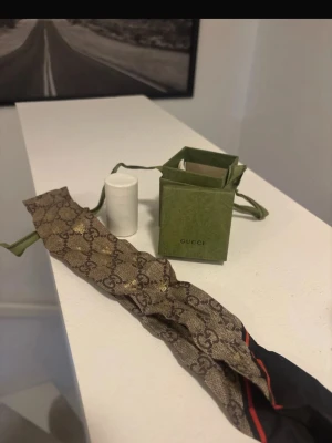 Gucci halsduk med GG-mönster - Snygg halsduk från Gucci i brunt med klassiskt GG-monogram och diskreta gulddetaljer. Halsduken har en smal, avlång form och är tillverkad i ett lyxigt tyg. Perfekt accessoar för att lyfta din outfit med en exklusiv touch.