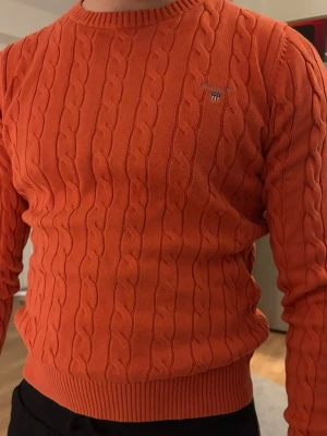 Orange kabelstickad tröja Gant - Säljer en orange kabelstickad tröja från Gant med rund halsringning och diskret logga broderad på bröstet. Tröjan har ribbade muddar vid ärmslut och nederkant, perfekt för dig som vill ha en färgstark och klassisk look.