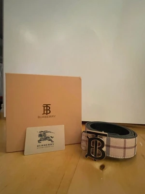 Beige rutigt bälte från Burberry - Stilrent bälte från Burberry i klassiskt beige rutigt mönster med en stor metallspänne med Burberrys logotyp. Bältet har detaljer i brunt och svart och är tillverkat i skinn och textil. Perfekt accessoar för att lyfta din outfit.
