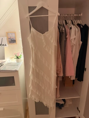 Vit volangklänning med smala axelband - Säljer en söt vit kortklänning med smala axelband och volangdetaljer som går diagonalt över hela klänningen. Klänningen har en lätt och luftig känsla, perfekt för varma dagar. Passar dig som gillar en feminin och trendig stil.