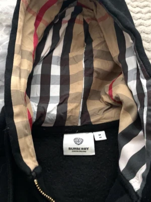 Svart Burberry hoodie med mönstrad huva - Snygg svart hoodie från Burberry med klassiskt rutigt foder i huvan i beige, svart, vitt och rött. Dragkedja framtill och tydlig Burberry-logga på insidan. Perfekt för dig som vill ha en stilren hoodie med lyxig känsla och ikoniskt mönster.