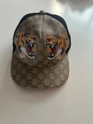 Gucci keps med tigertryck - Säljer en beige Gucci keps med klassiskt GG-mönster och två broderade tigrar framtill. Kepsen har svart mesh baktill och justerbart band med rödgrön detalj. Materialet är en mix av bomull och polyester, perfekt för en trendig streetwear-look.