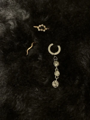 Silvrigt örhänge med tre stenar - Säljer en silvrig Navelpiercing med tre hängande, runda klara stenar som ger en glittrig och trendig look. 2 guldiga ringar (inte äkta)