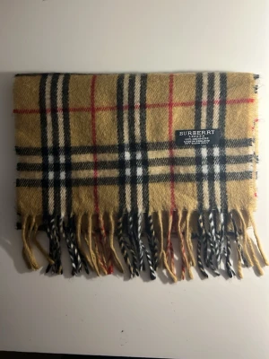 Beige rutig halsduk från Burberry - Klassisk beige halsduk från Burberry med det ikoniska rutmönstret i svart, vitt och rött. Halsduken har tvinnade fransar i ändarna och är tillverkad i mjukt material som känns lyxigt och stilrent. Perfekt accessoar för att lyfta vilken outfit som helst.