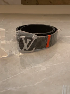 Svart bälte med LV-spänne - Snyggt svart bälte med klassiskt LV-spänne i silverfärg. Bältet har en diskret mönstrad yta och en orange detalj. Perfekt accessoar för att lyfta din outfit.