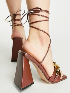 Bruna sandaler med blockklack och kedja - Trendiga bruna sandaler med höga blockklackar och tunna remmar som snöras runt ankeln. Framtill finns en bred rem med en stor guldfärgad kedjedetalj. Skorna har fyrkantig tå och är tillverkade i ett läderliknande material.