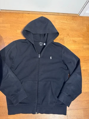 Svart hoodie från Polo Ralph Lauren - Klassisk svart hoodie från Polo Ralph Lauren med dragkedja framtill och den ikoniska vita logon broderad på bröstet. Hoodien har känguruficka och huva, tillverkad i mjukt bomullsmaterial. Perfekt för en avslappnad och stilren look.