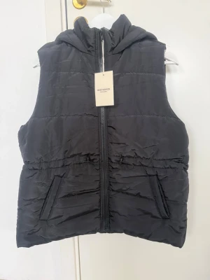 Svart dunväst med huva från New Fashion - Snygg svart dunväst från New Fashion med dragkedja framtill, två fickor och justerbar midja med snörning. Västen har en mysig huva och quiltad design som ger en modern look. Perfekt för lager-på-lager och enkel att matcha till olika outfits.