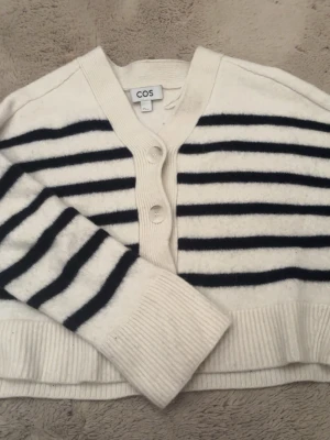 Vit och svart randig kofta från COS - Vit kofta från COS med svarta ränder. Tröjan har lång ärm och ribbade muddar vid ärmslut och nederkant. Perfekt för en stilren och avslappnad look.