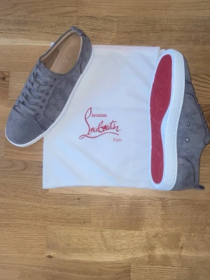 Gråa Louboutin sneakers i mocka - Snygga gråa sneakers från Christian Louboutin i mjuk mocka med klassisk röd sula och vita kanter. Lågt skaft och stilren design med matchande grå skosnören. Perfekta för dig som vill ha något exklusivt och trendigt i garderoben.