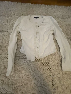 Vit kofta från Tommy Hilfiger - Klassisk vit kofta från Tommy Hilfiger med rund halsringning och knappar framtill. Liten broderad logga på bröstet. Tillverkad i mjukt material och har långa ärmar samt ribbade muddar vid ärmslut och nederkant.