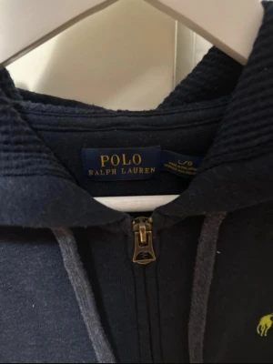Zip hoodie Polo Ralph Lauren - Mörkblå hoodie från Polo Ralph Lauren med dragkedja framtill och klassisk huva. Ikoniska gröna loggan broderad på bröstet. Tillverkad i mjukt bomullsmaterial som känns skönt mot huden. Perfekt för en avslappnad och stilren look.